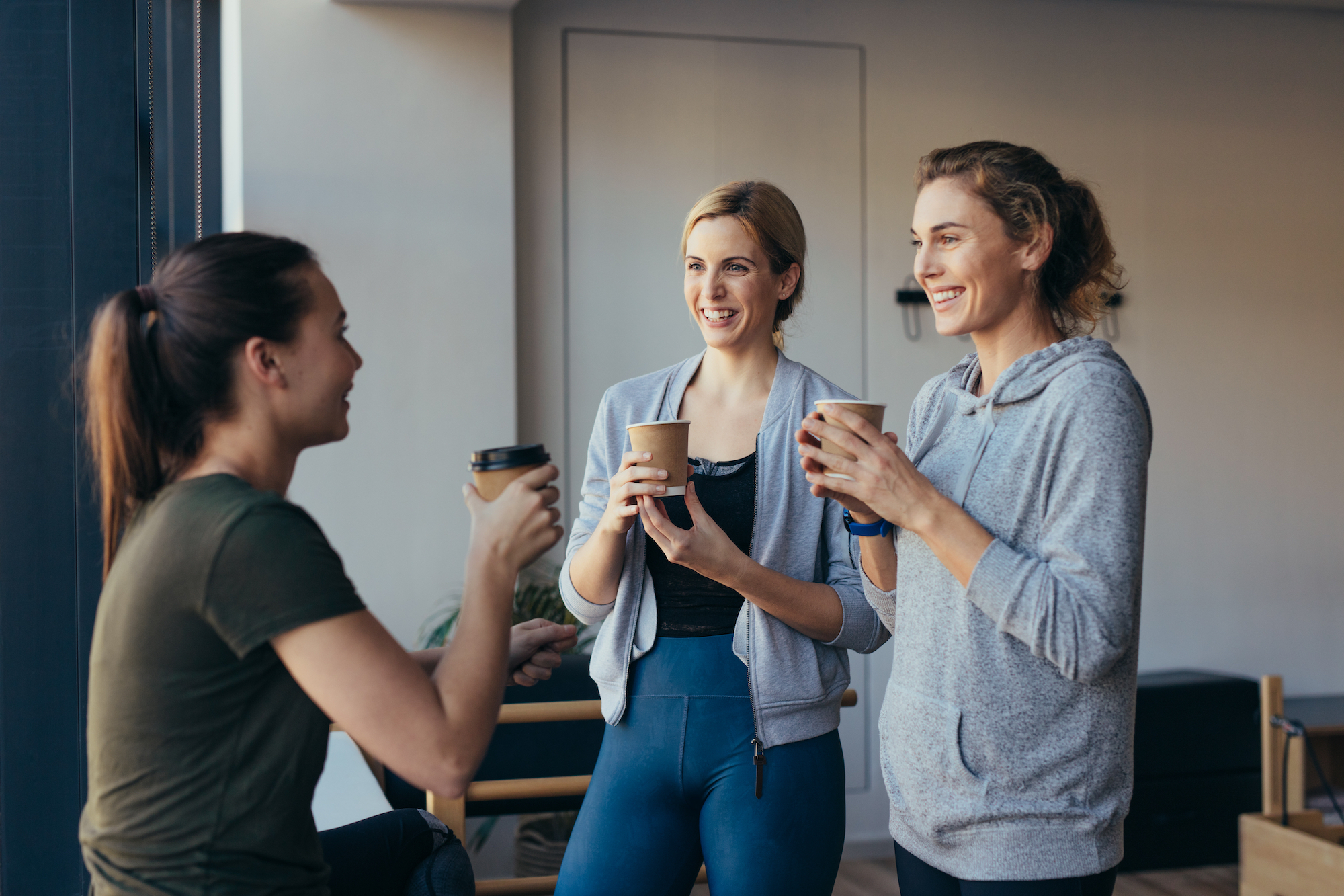 Three women in athletic wear holding coffee cups in a modern cafe