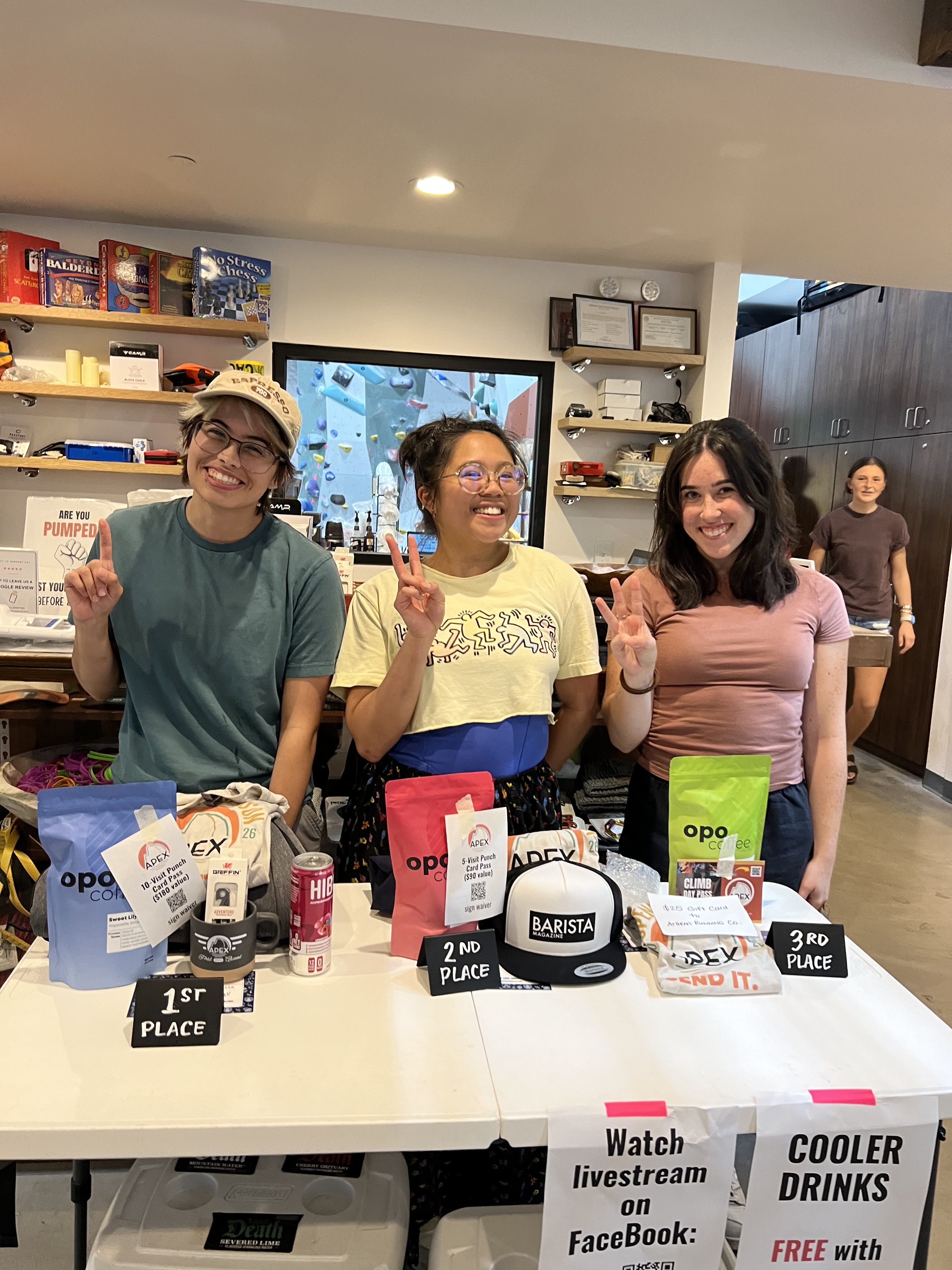 Three people behind the prize table holding up fingers and smiling at the end of the Bouldering Baristas throwdown
